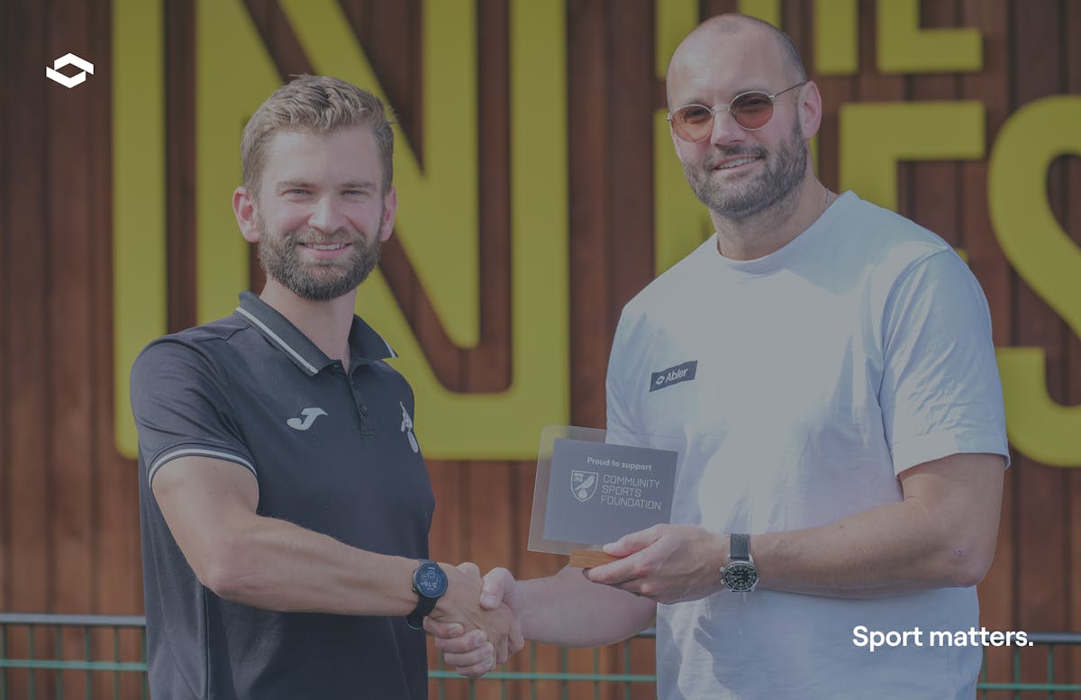 Two men shaking hands in front of Norwich City FC signage at Carrow Road stadium. On the left, Freddie Powell-Tuck wearing a dark Norwich City polo shirt, and on the right, Markus Maute in a white Abler t-shirt, holding a Community Sports Foundation folder while completing their partnership handshake.