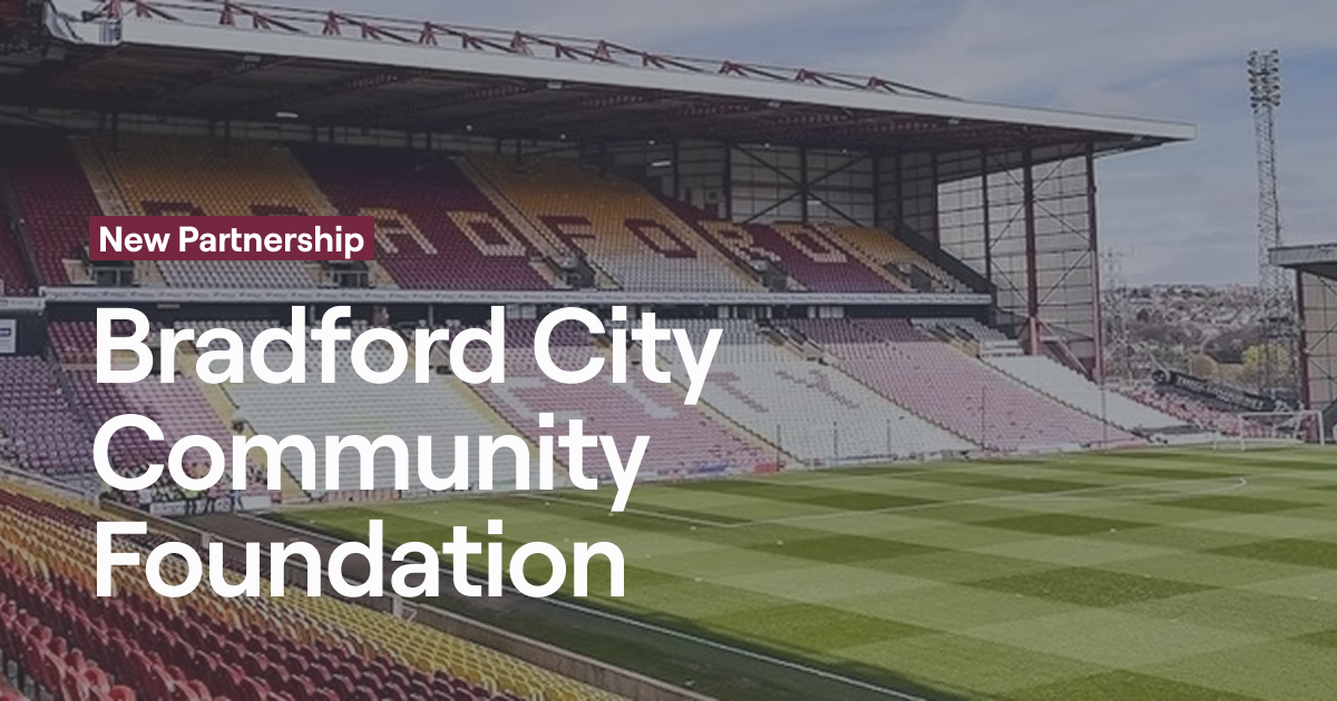 The main stand at Bradford City FC's stadium, Valley Parade, showing the distinctive burgundy and amber seats spelling out "BCFC" with the pitch visible in the foreground. Text overlay reads "New Partnership: Bradford City Community Foundation" in white text against the stadium backdrop.