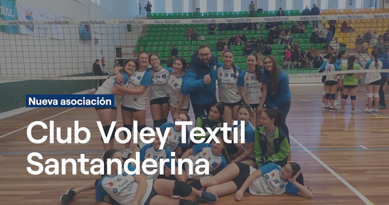 Equipo femenino del Club Voley Textil Santanderina posando en grupo en la cancha con sus entrenadoras, vistiendo uniformes blancos y azules, con gradas verdes y amarillas al fondo