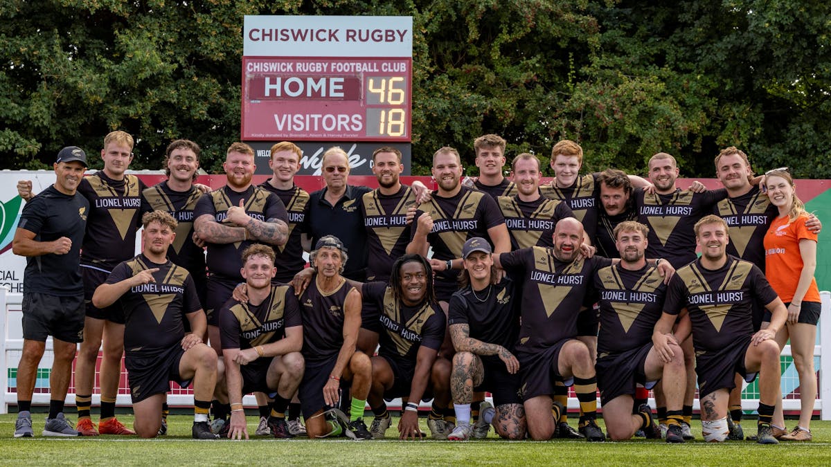 All Golds Rugby League senior team posed together on the pitch at Chiswick Rugby Football Club. The team of approximately 25 players wearing black and gold Lionel Hurst sponsored jerseys are arranged in two rows, with the scoreboard showing "Home 46, Visitors 18" visible in the background.