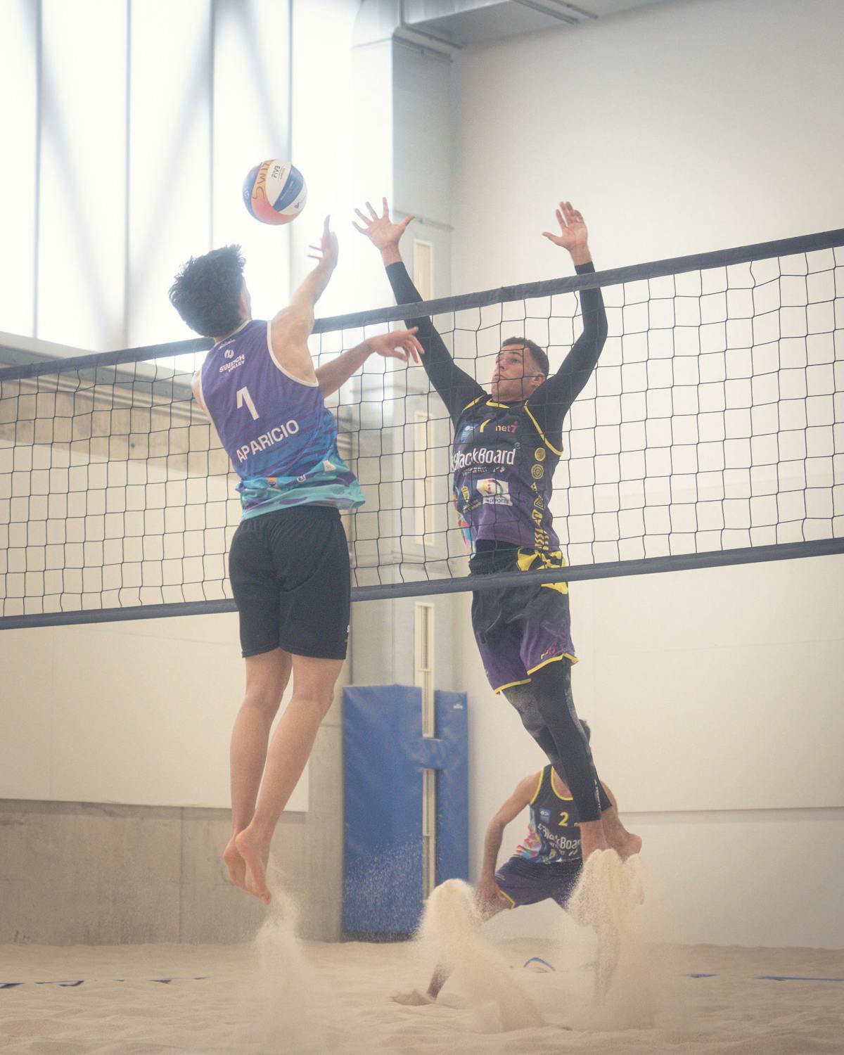 Two players competing at the net during an indoor beach volleyball match, one wearing Switch Volley uniform