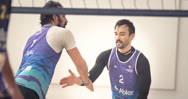 Two Switch Volley players in purple and blue uniforms high-fiving during an indoor beach volleyball match