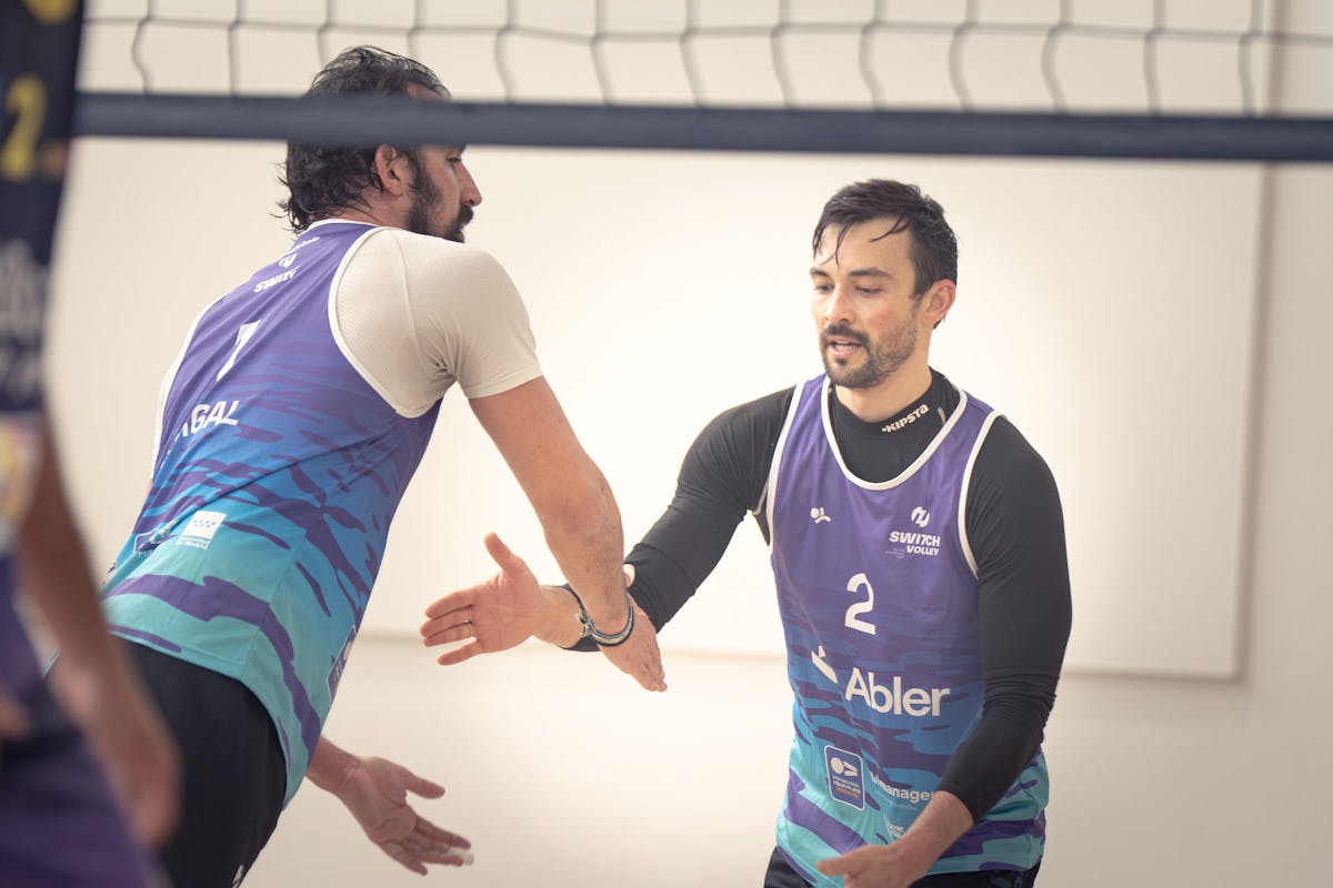 Two Switch Volley players in purple and blue uniforms high-fiving during an indoor beach volleyball match
