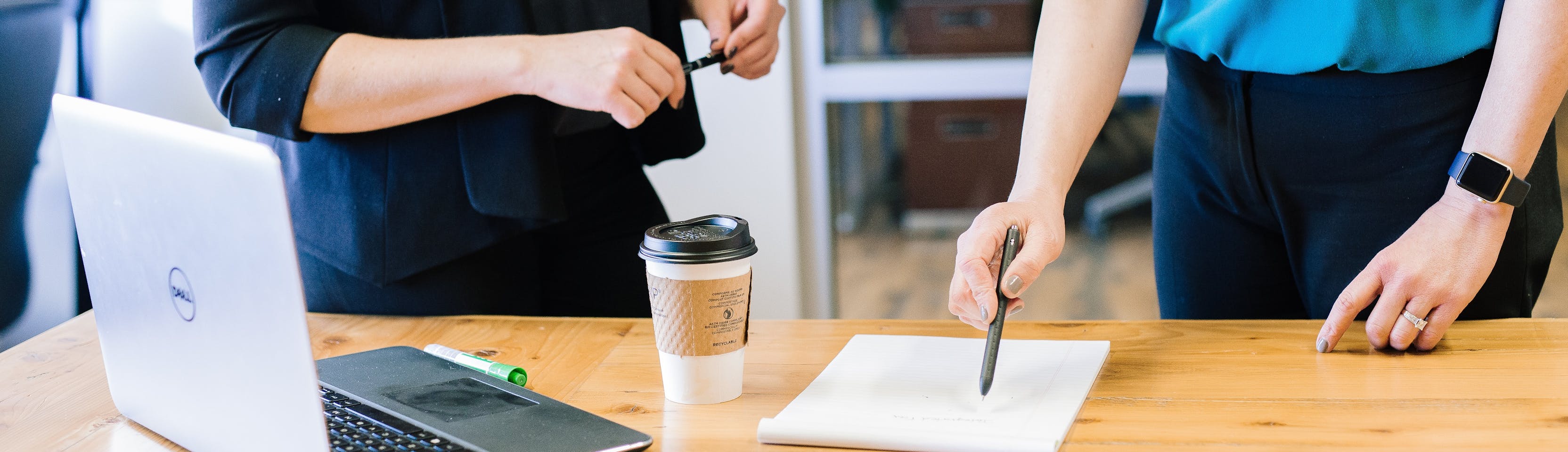 The hands of two office workers holding pens and pointing at a notepad lying next to an open laptop