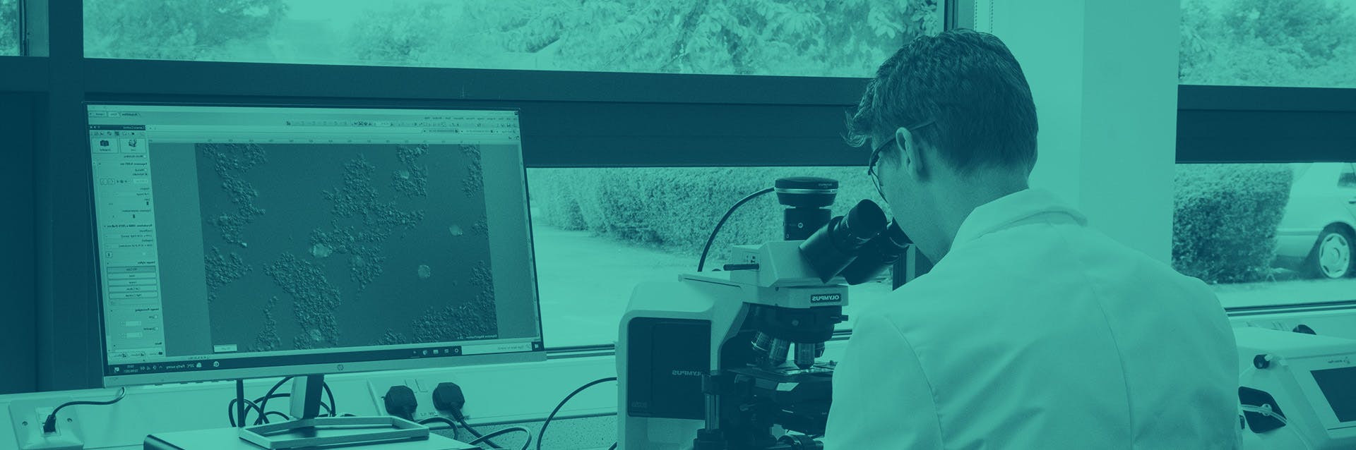 A scientist in a lab coat sits at a desk, looking into a microscope.