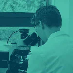 A scientist in a lab coat sits at a desk, looking into a microscope.