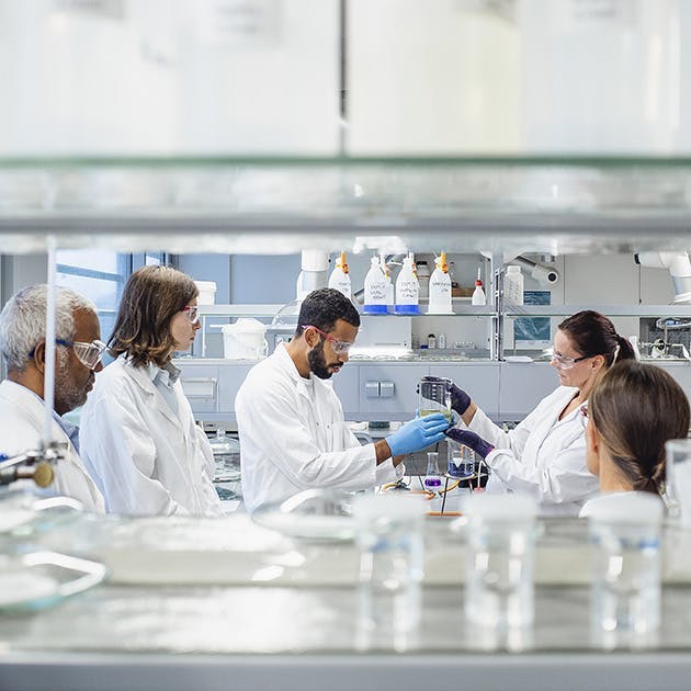 Scientists gather around to examine a large glass beaker that two of the scientists are holding. All are experts wearing white lab coats, gloves and safety glasses and are in a lab.