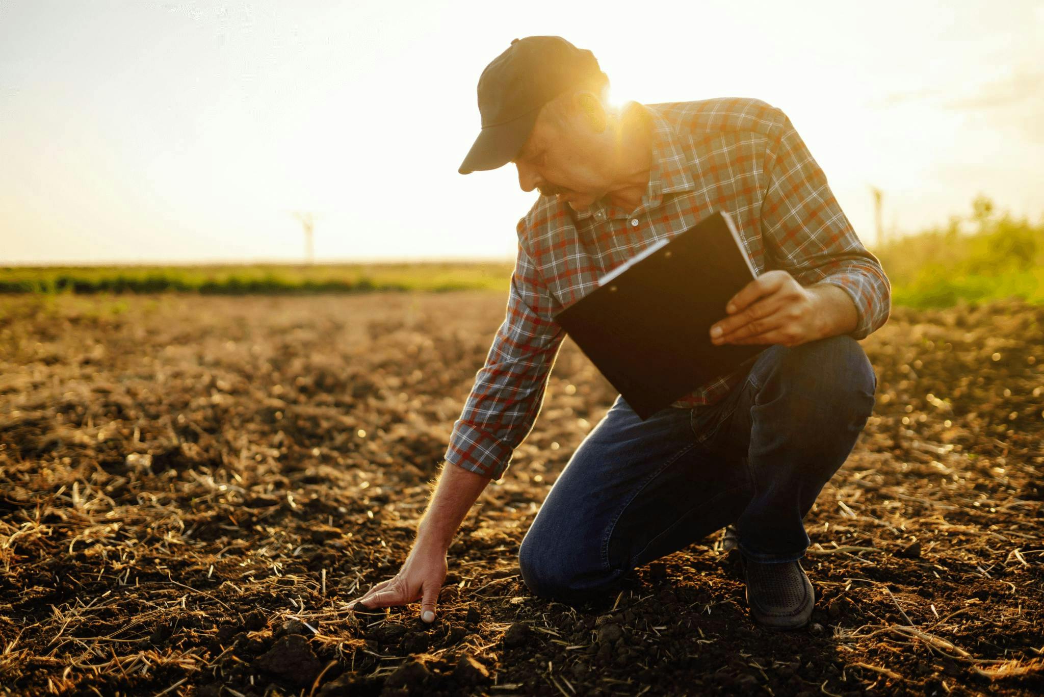 A farmer holding a clip board kneels on one knee in a field, one hand examining the dark brown soil.