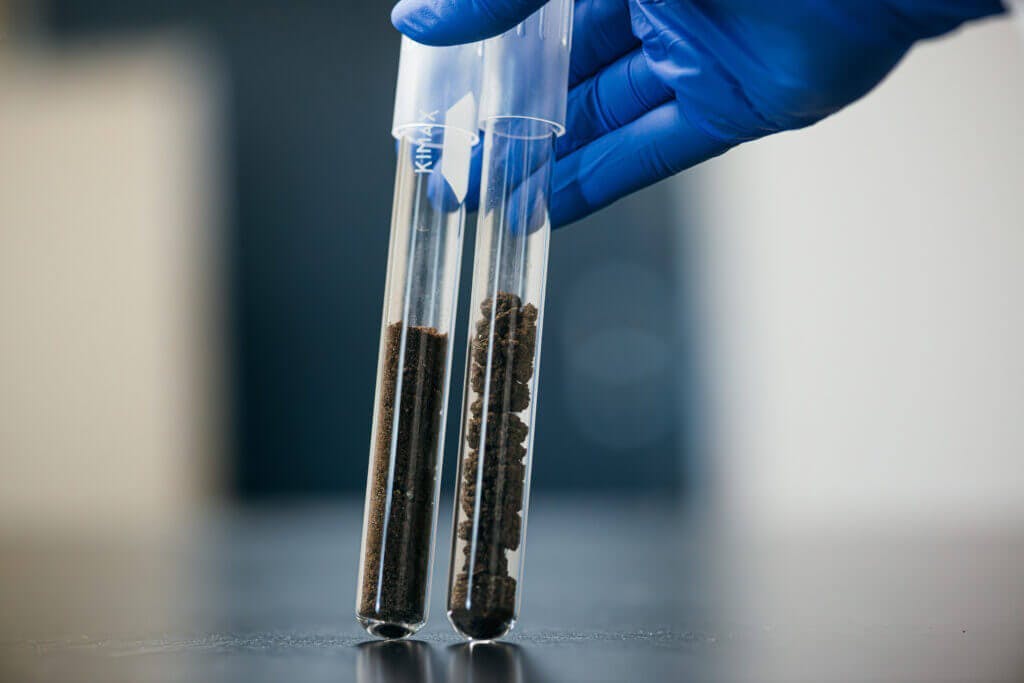 Two clear test tubes being held side by side by a blue-gloved hand. There is brown soil in each test tube.