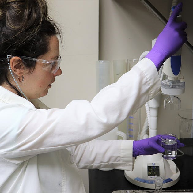 A person in a white lab coat and purple gloves is holding a piece of lab equipment in one hand and a beaker of clear liquid in the other.