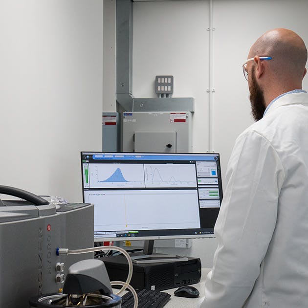 A scientist reviews a computer screen in a laboratory. He is wearing a white lab coat and protective glasses.