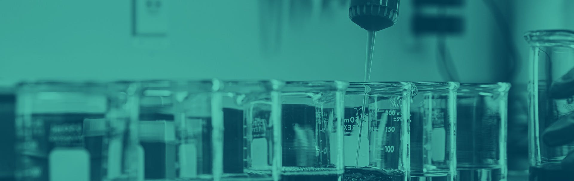 Scientific glass beakers are bunched together on a desk, with a liquid being added into one.