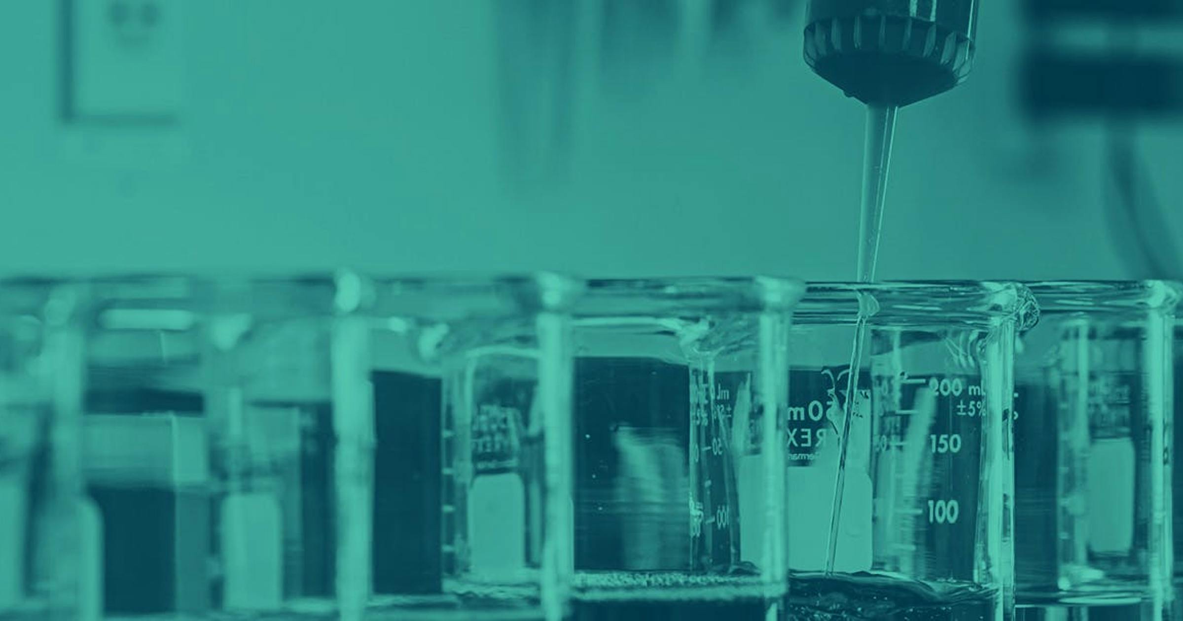 Scientific glass beakers are bunched together on a desk, with a liquid being added into one.