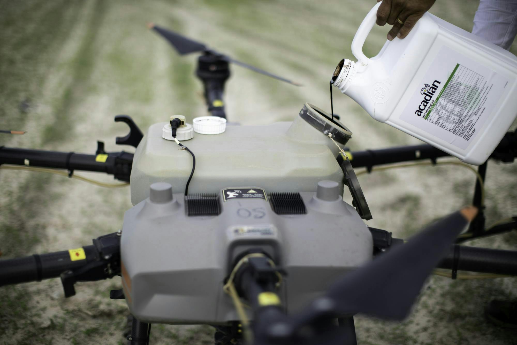 A large drone is on the ground. It has a large tank that’s being filled with a jug of product marked with the Acadian branding.