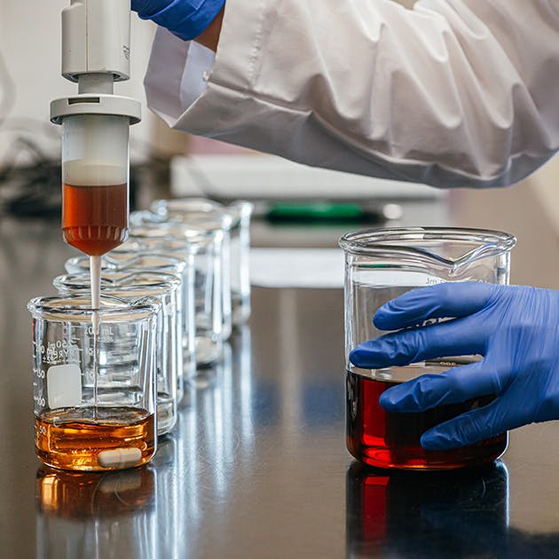A close up of a person in a white lab coat emptying a syringe of amber liquid into a beaker. 