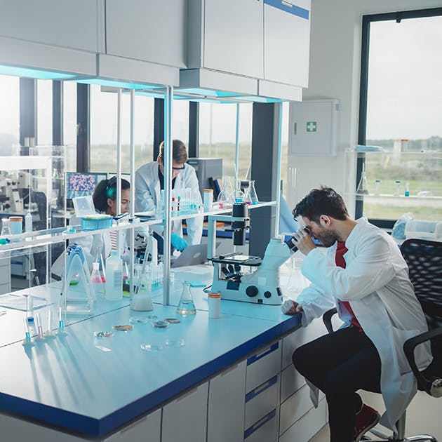 A scientist sits at a lab bench gazing into a microscope. He has beakers and tools beside him, and two more scientists are working on a laptop across from him.