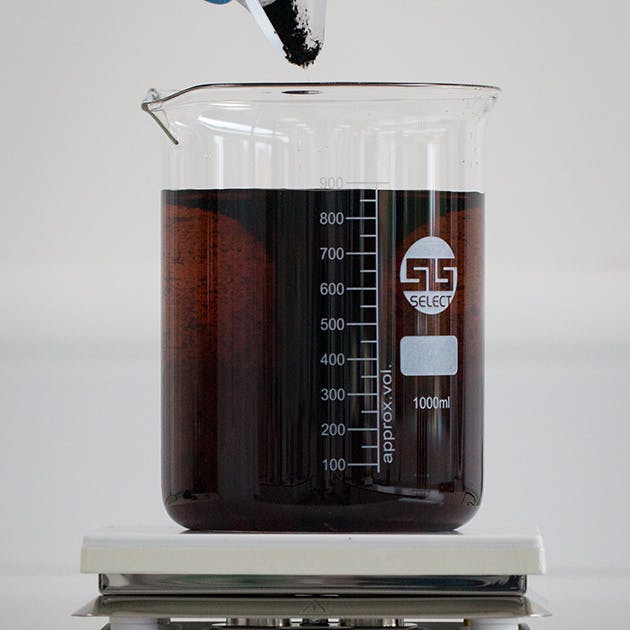 A large beaker of water sits on a white scientific scale. The water is brown and murky, and a soil sample is being carefully added to the water.