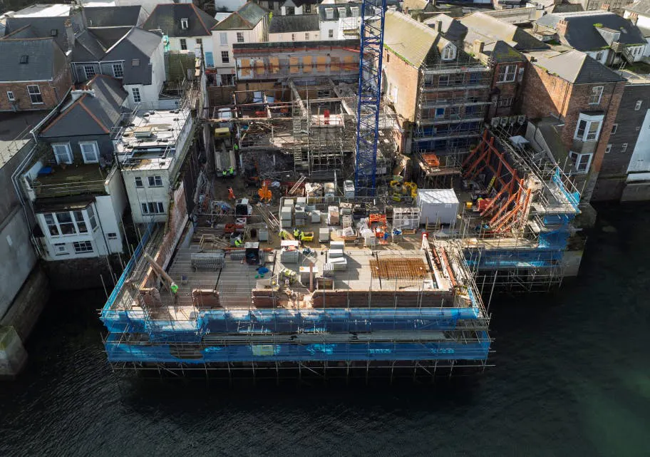 View of the build fronting the water. The structure continues to rise, with brickwork, scaffold lifts, and block laying underway across the podium, assisted by the tower crane.