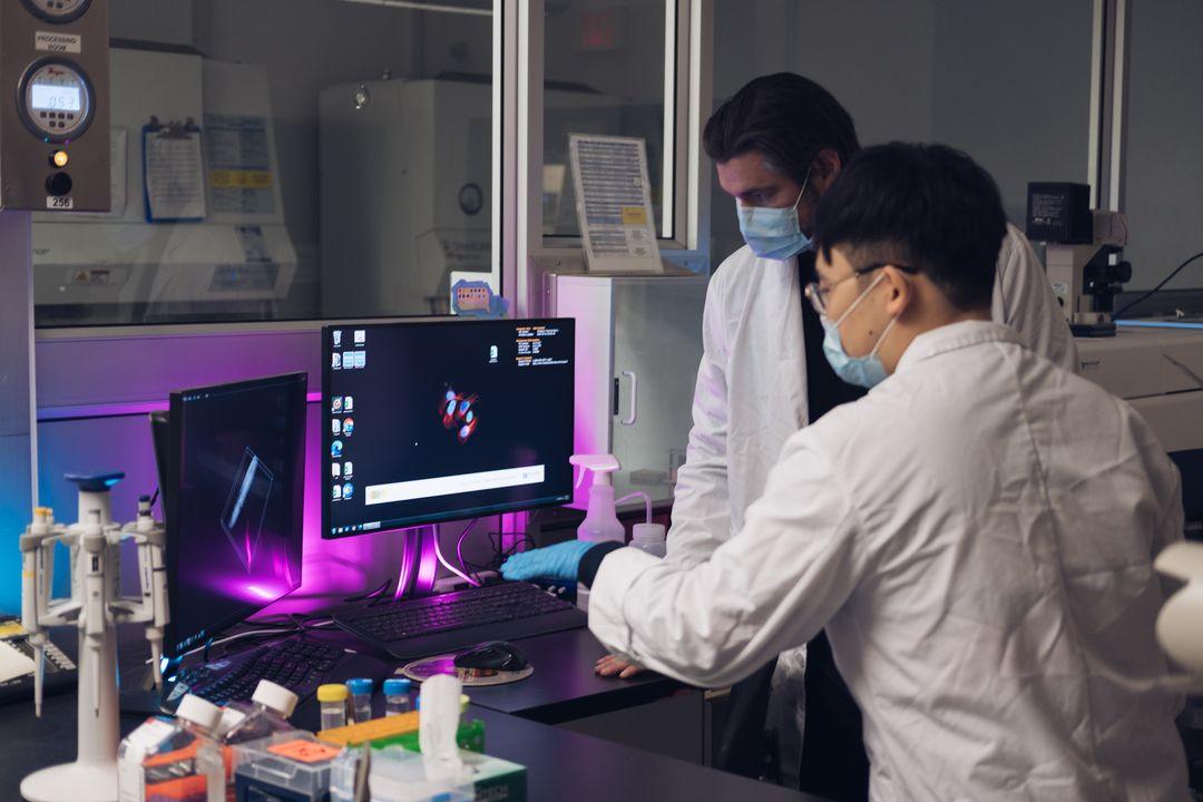 Two researchers in lab coats collaborate at computer screens in a laboratory setting.