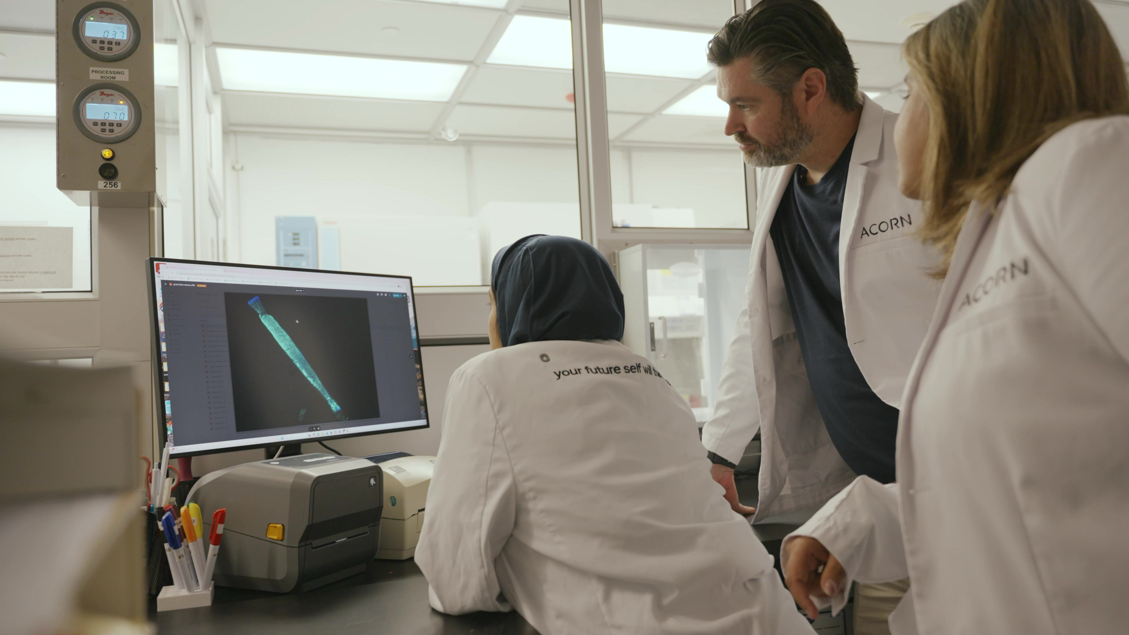 Two masked professionals in lab coats analyze data on computer screens in a research environment.