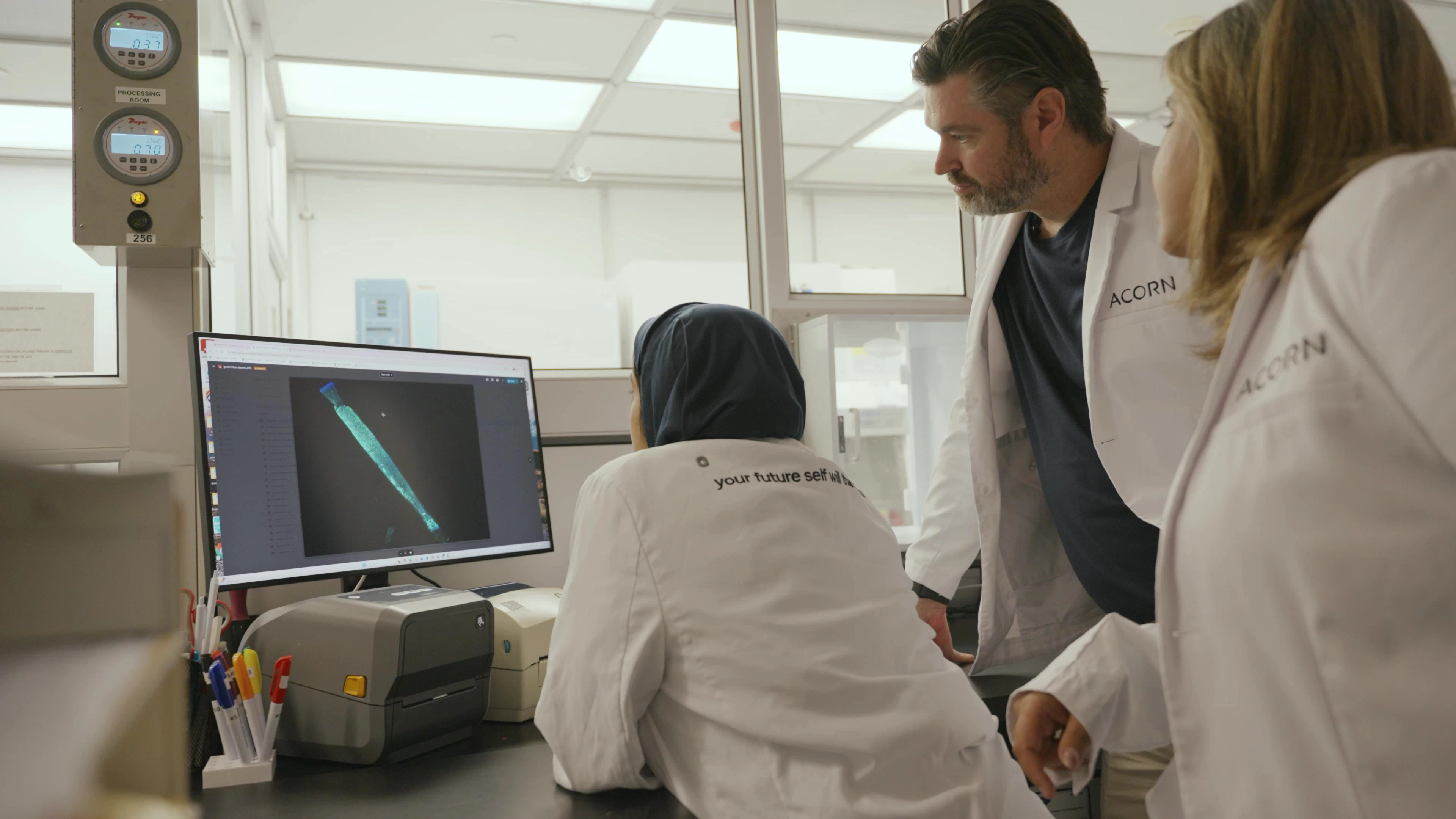 Two masked professionals in lab coats analyze data on computer screens in a research environment.