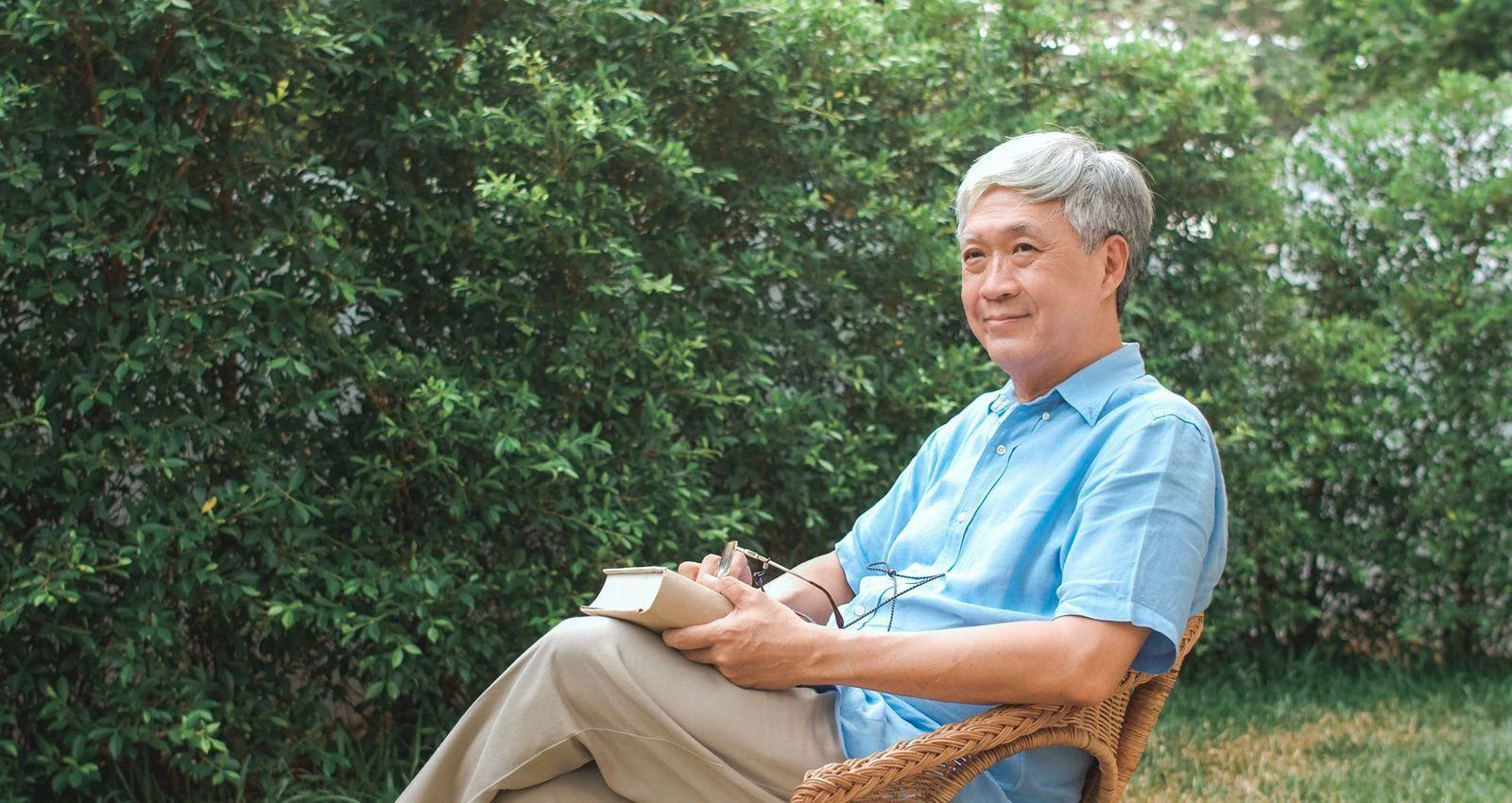 A senior man comfortably seated in a wicker chair, focused on reading a book in a warm, inviting space.