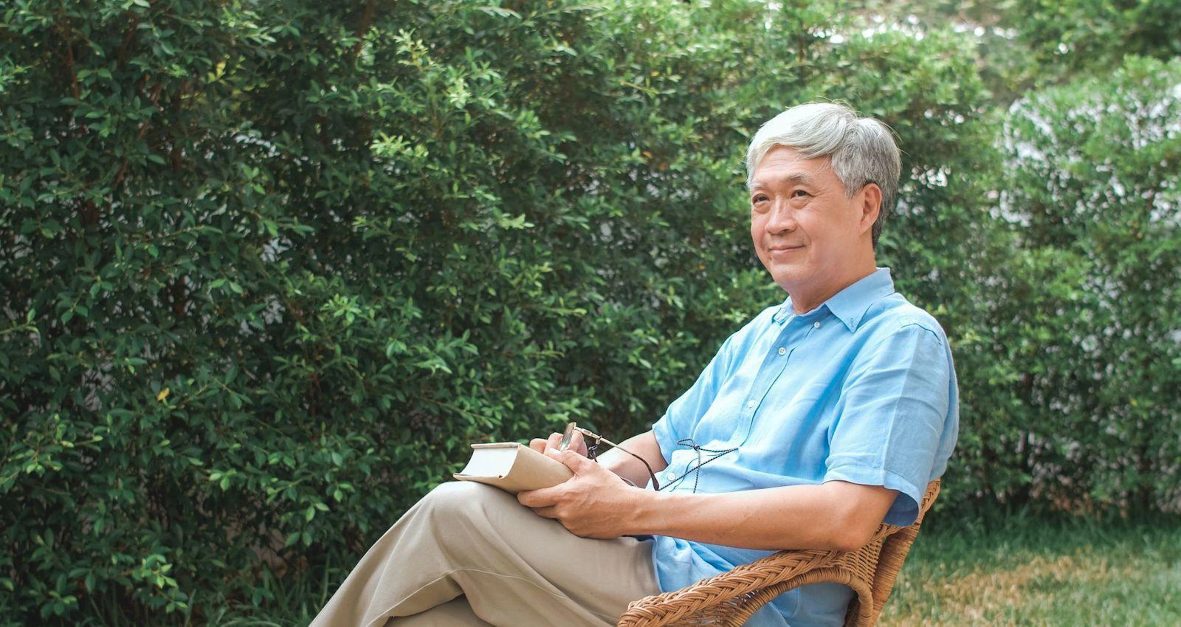 A senior man comfortably seated in a wicker chair, focused on reading a book in a warm, inviting space.