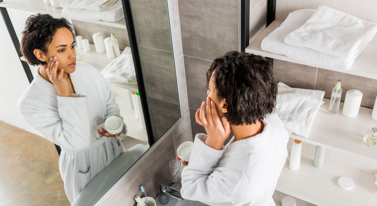 A woman in a bathrobe examines her reflection in a mirror, focusing on her facial features.