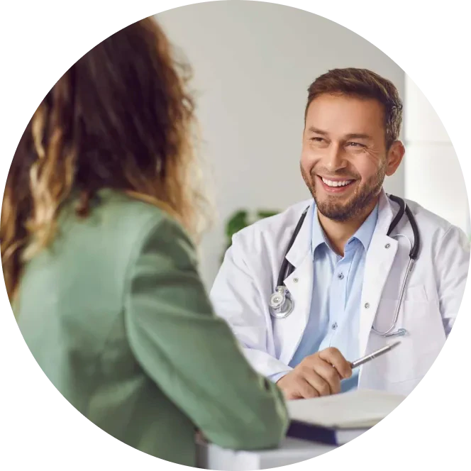 A doctor in a white coat with a stethoscope smiles and talks with a patient sitting across the desk in an office setting.