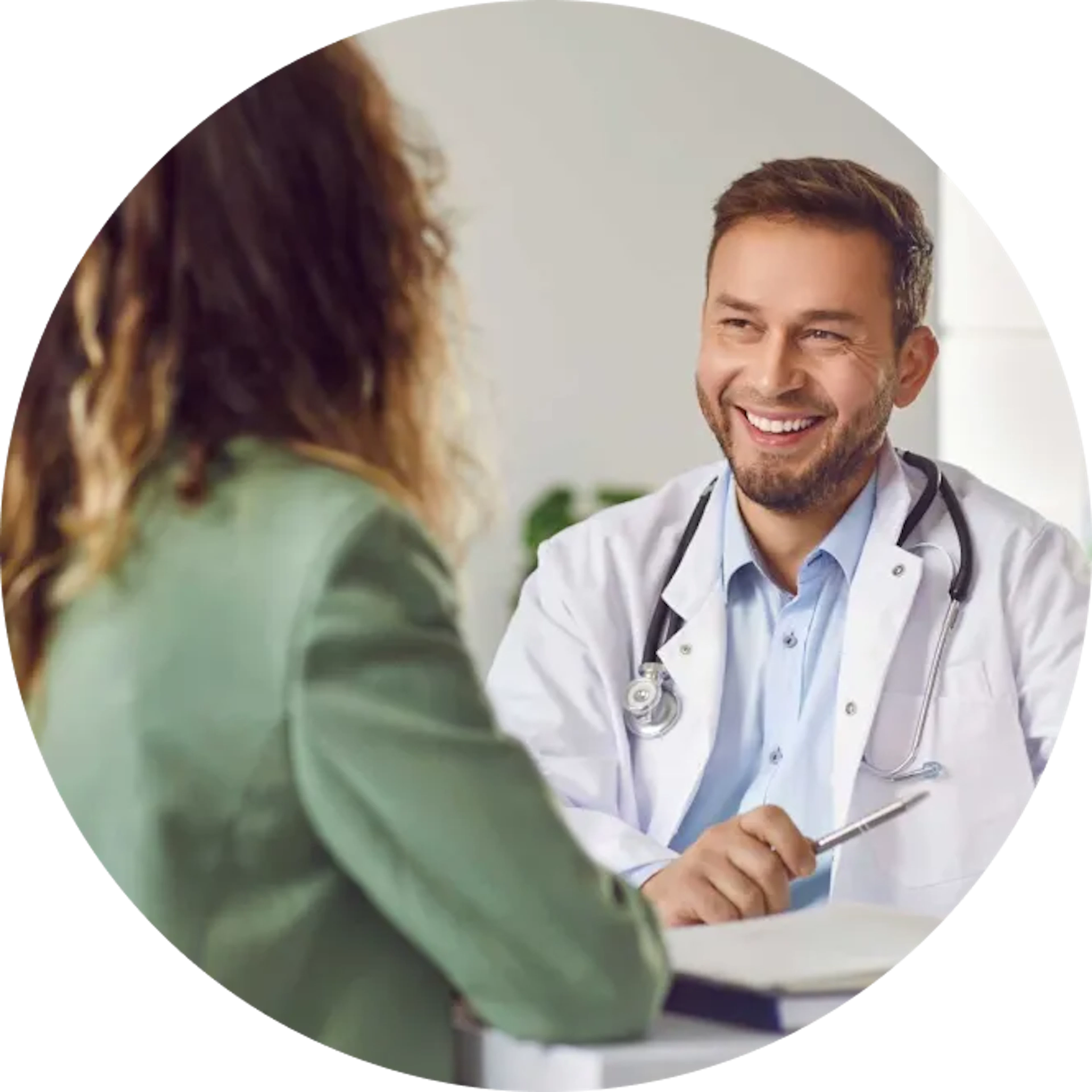 A doctor in a white coat with a stethoscope smiles and talks with a patient sitting across the desk in an office setting.