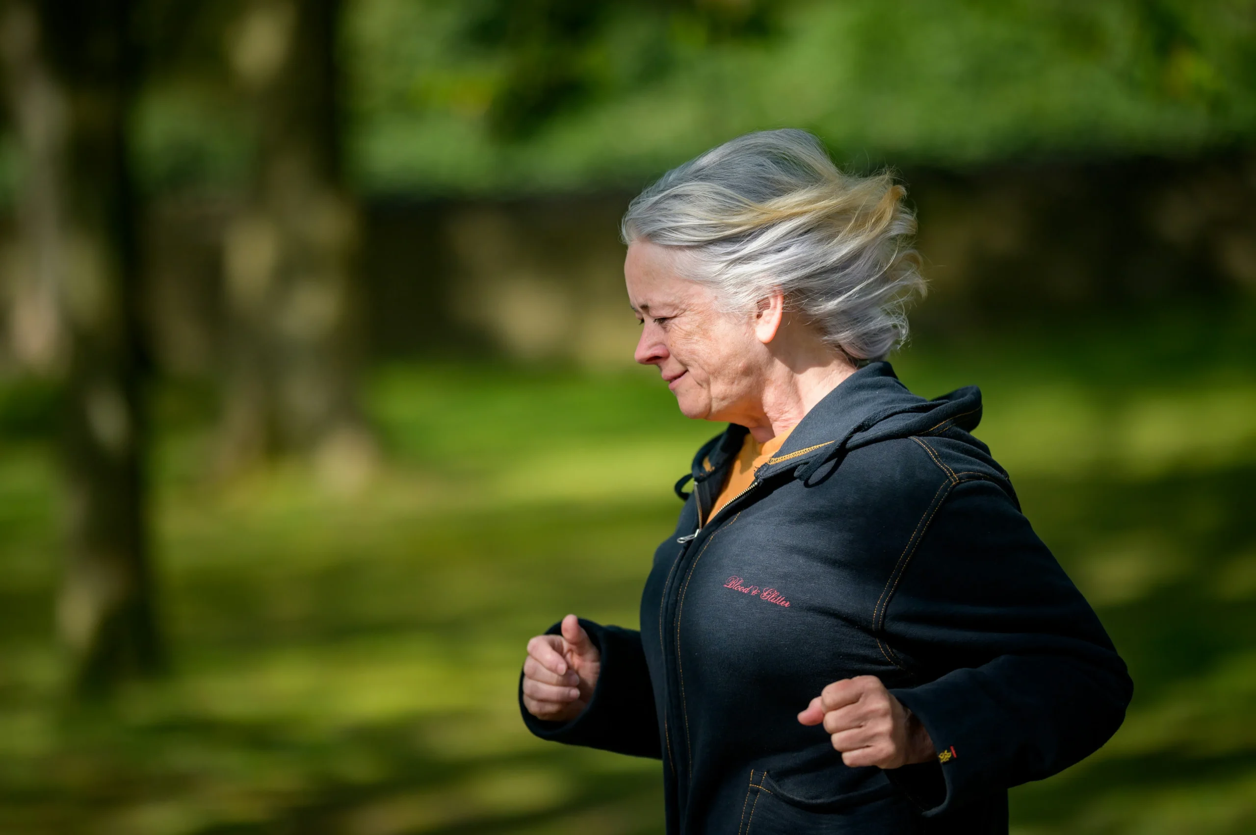Older woman with gray hair jogs outdoors in a park, wearing a dark zip-up jacket. Sunlight filters through trees in the background.