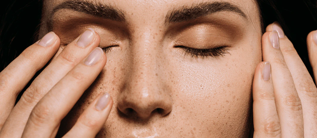 A woman with freckles gently touches her eyes, showcasing a thoughtful expression on her face.