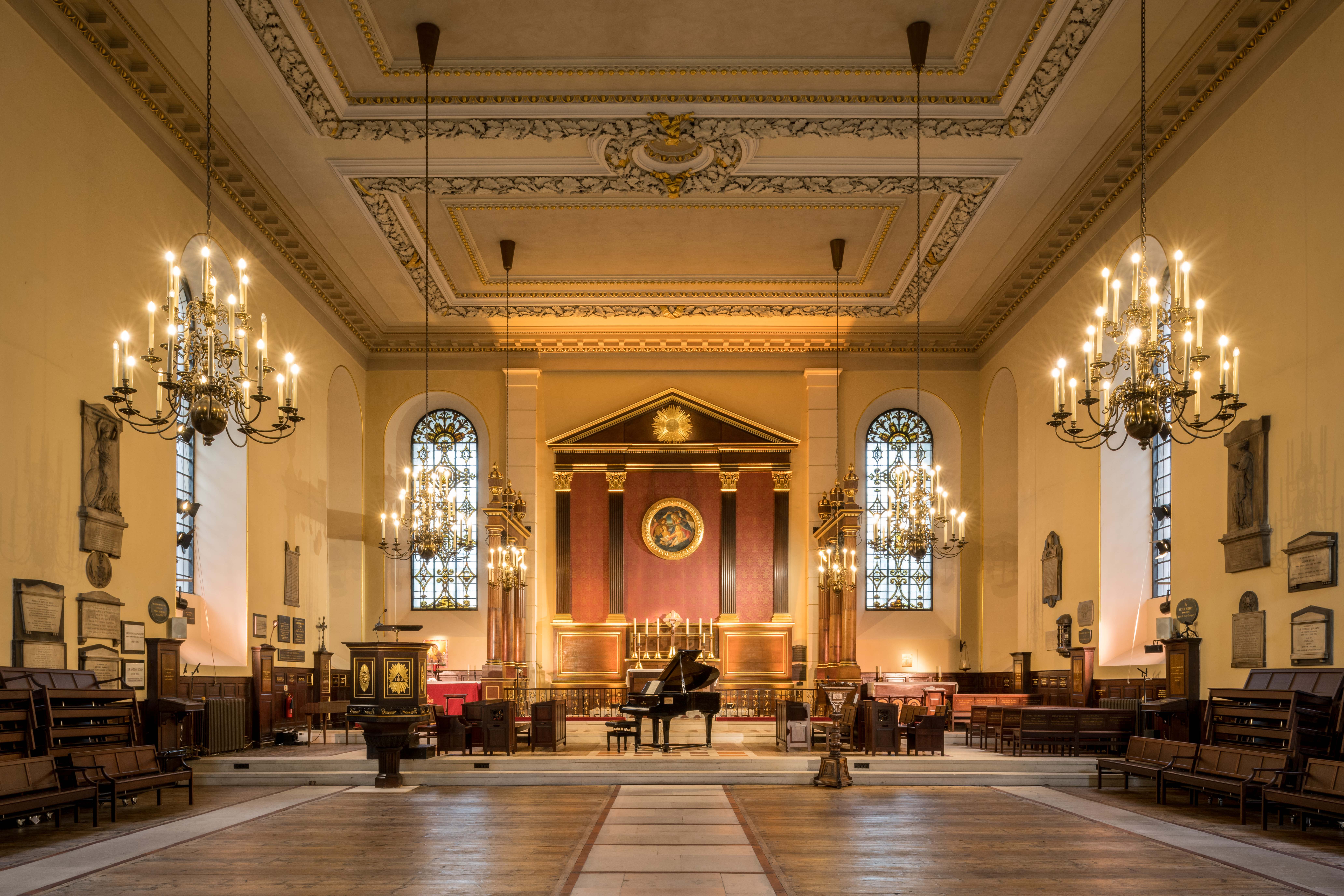 A view of the church with the pews stacked on each side. There is a grand piano in the centre in front of the altar. The sanctuary and altar are brightly lit.