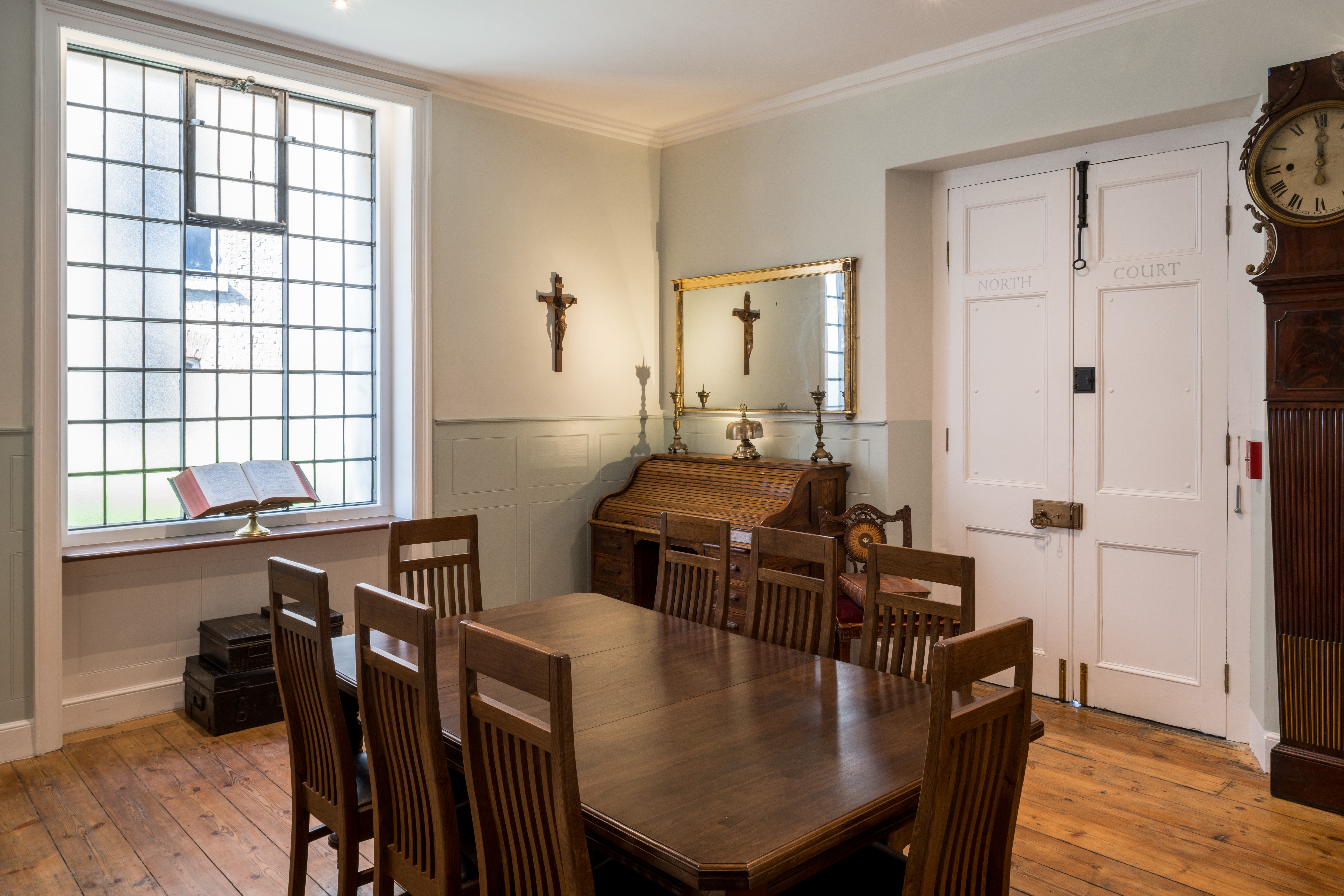 A large table surrounded by eight chairs. In the background, a desk and mirror. To the right, a door marked North Court. To the left, a large window with an open Bible on the sill.