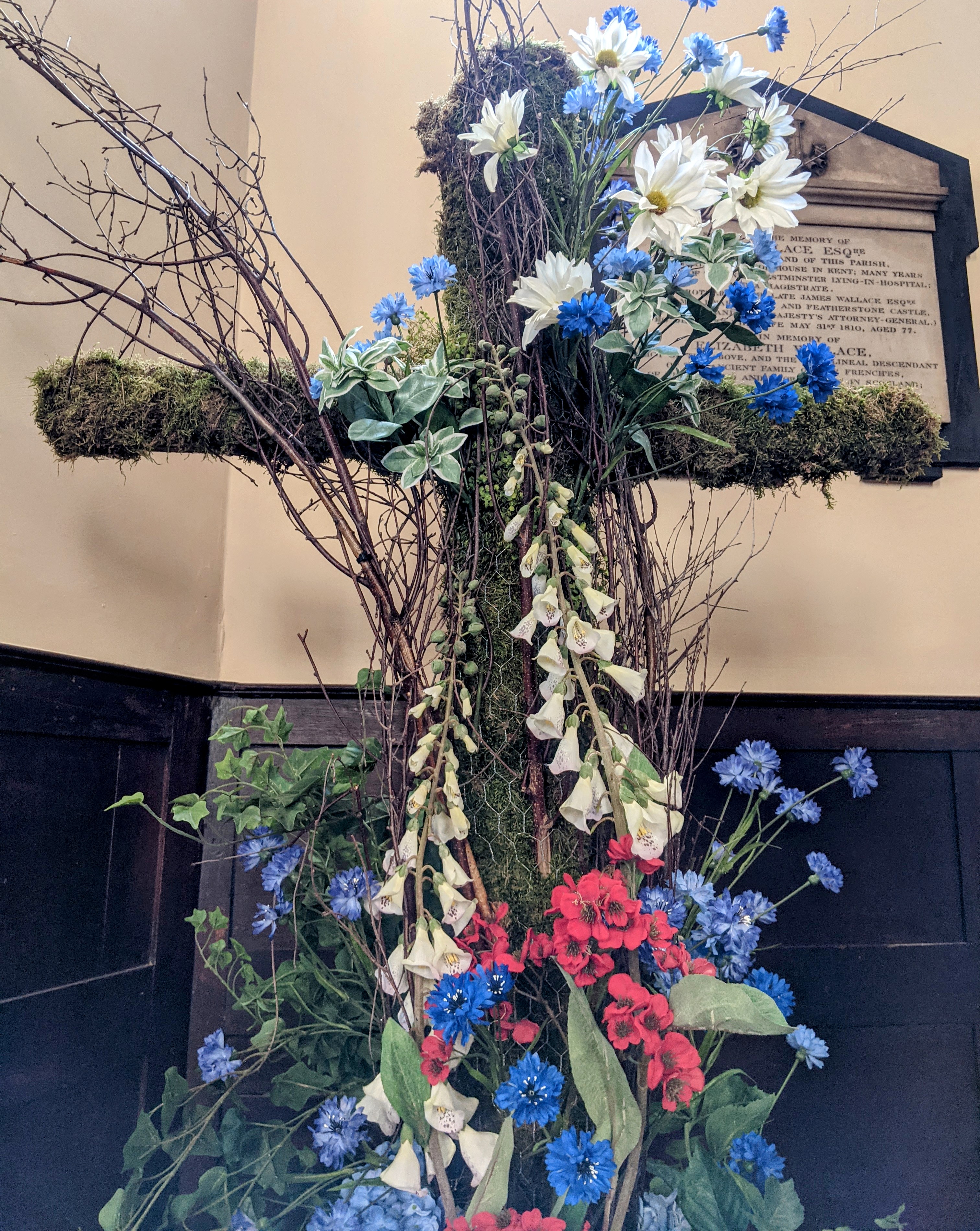 A cross made of chicken wire and moss is decorated with flowers and branches to create a floral arrangement.
