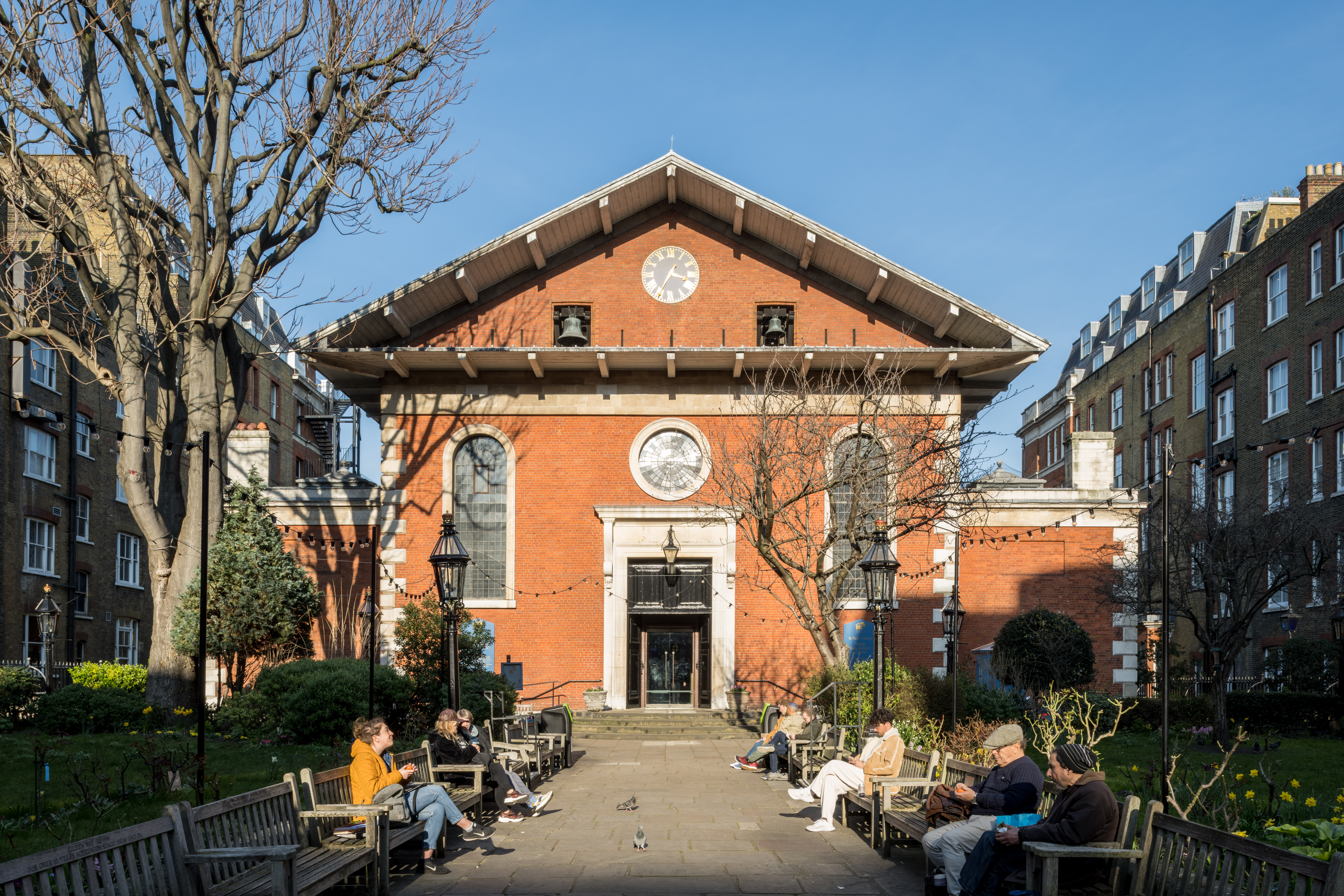 A view of the main entrance of the church in winter with a blue sky and people sat on the benches.