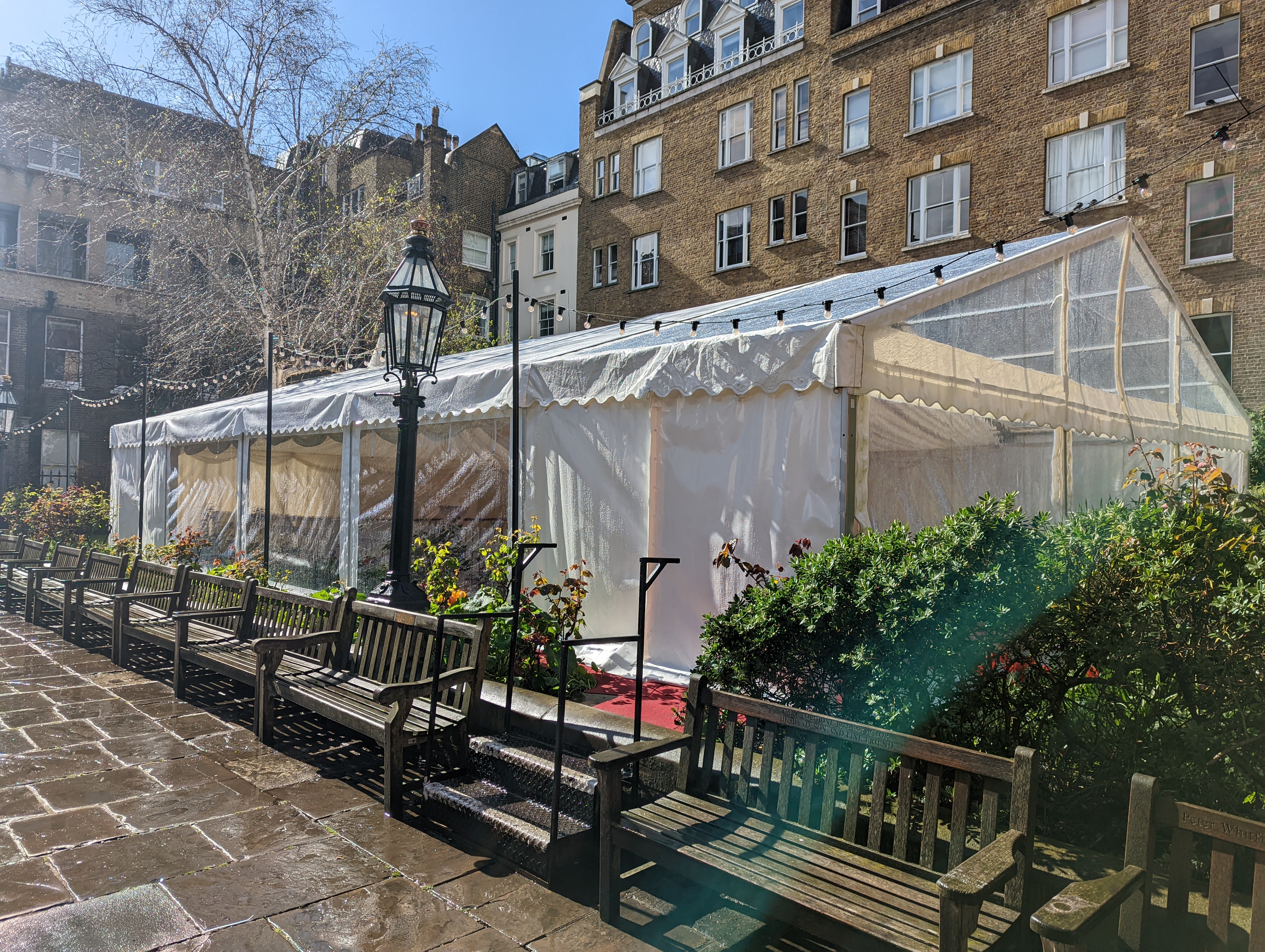 A large white marquee spans the length of the garden. There are steps up to it and benches along the pathway by it. There is a lamppost in the foreground and a silver birch tree in the background.