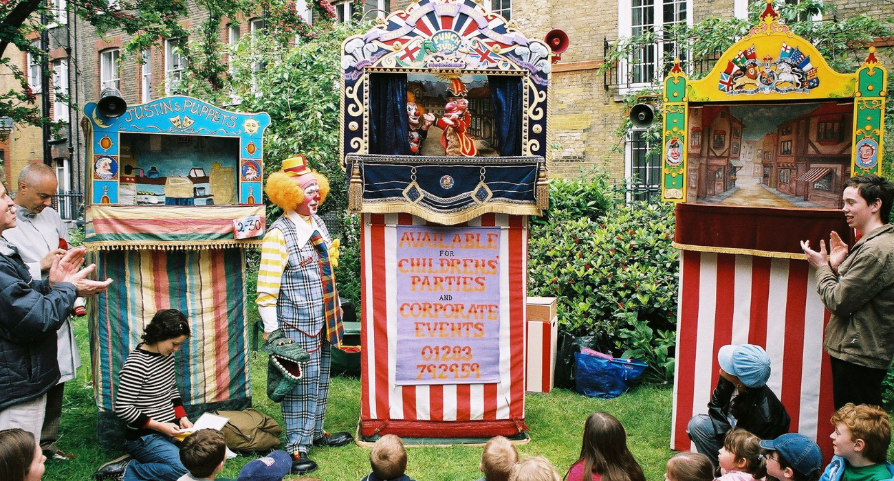 Outside a collection of children sit in front of a brightly coloured box with a puppet theatre on top. Within the puppets of Punch and Judy argue. Next to the puppet theatre stands a man dressed in an oversized suit, with bright curly hair and a face painted like that of a clown, he is smiling at the chaos. The sun is out and the grass underneath is fresh. Others stand around the edge applauding. 