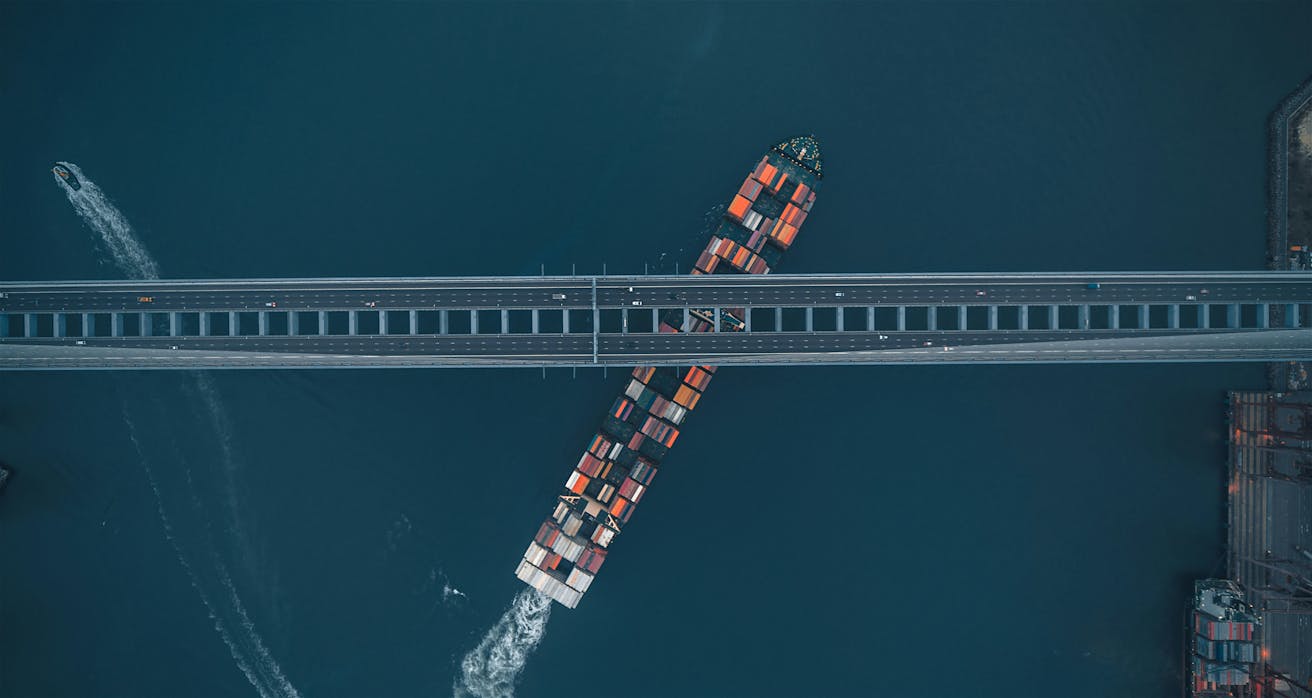 Container ship passing beneath a busy highway bridge, symbolizing global logistics and wholesale distribution of electronic components