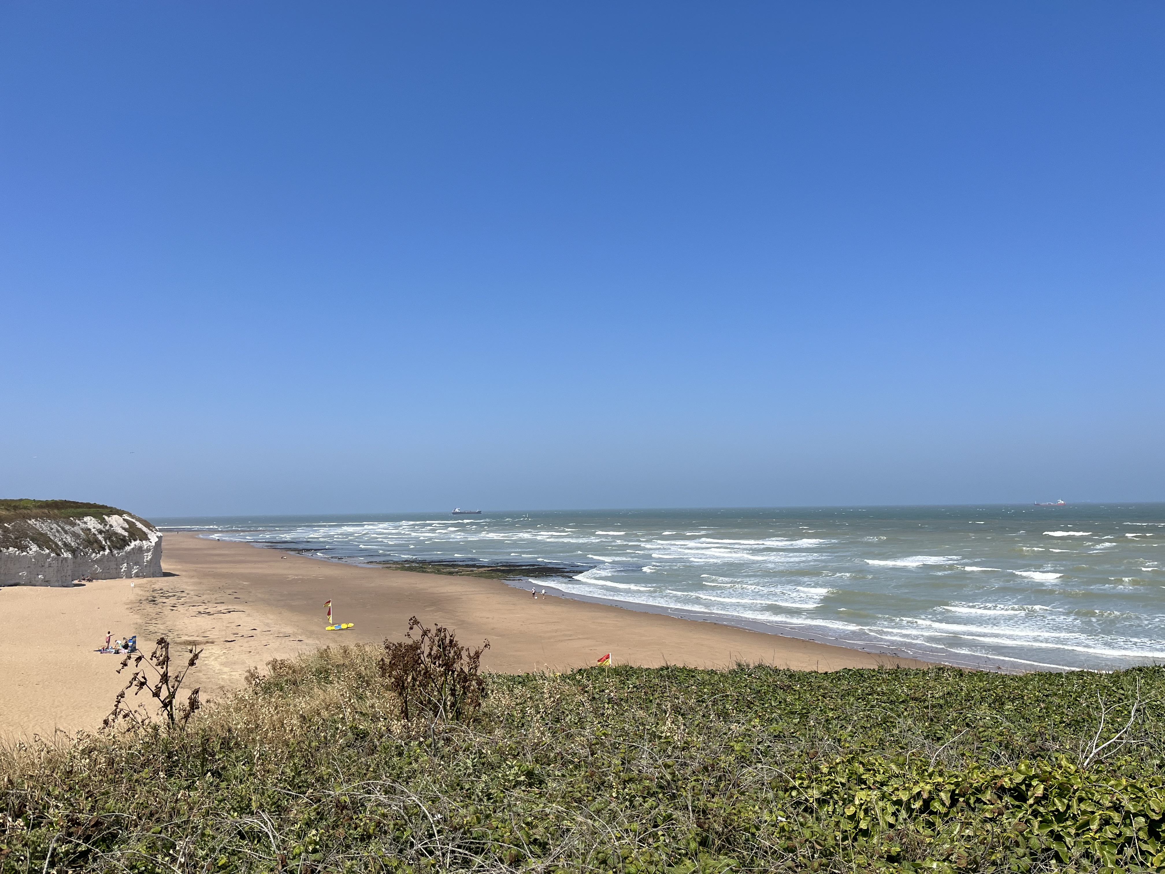 A photo of a sandy beach under clear blue skies. There is some scrub in the foreground, white cliffs in the distance, and some people on the sand.