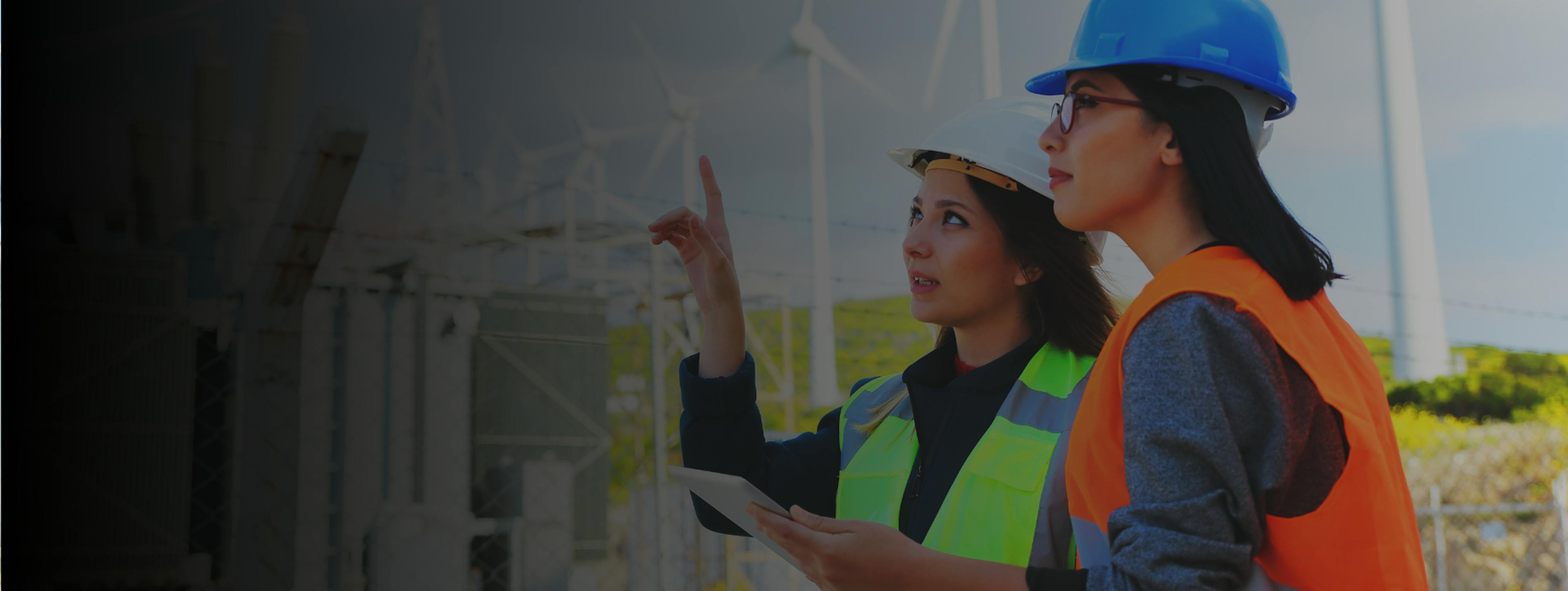 Two women wearing hi-vis vests and hard hats, standing in front of wind towers