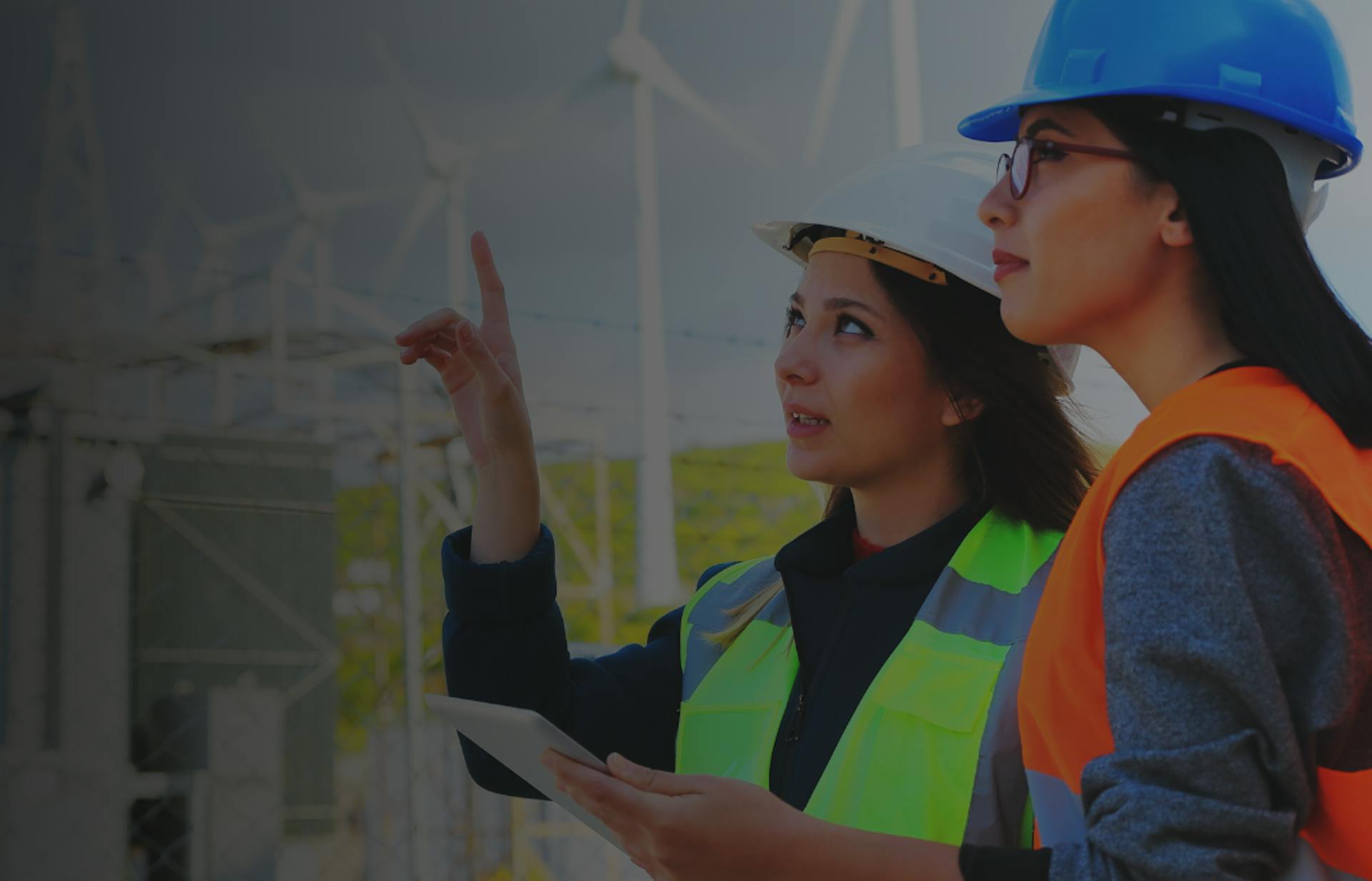 Two women wearing hi-vis vests and hard hats, standing in front of wind towers