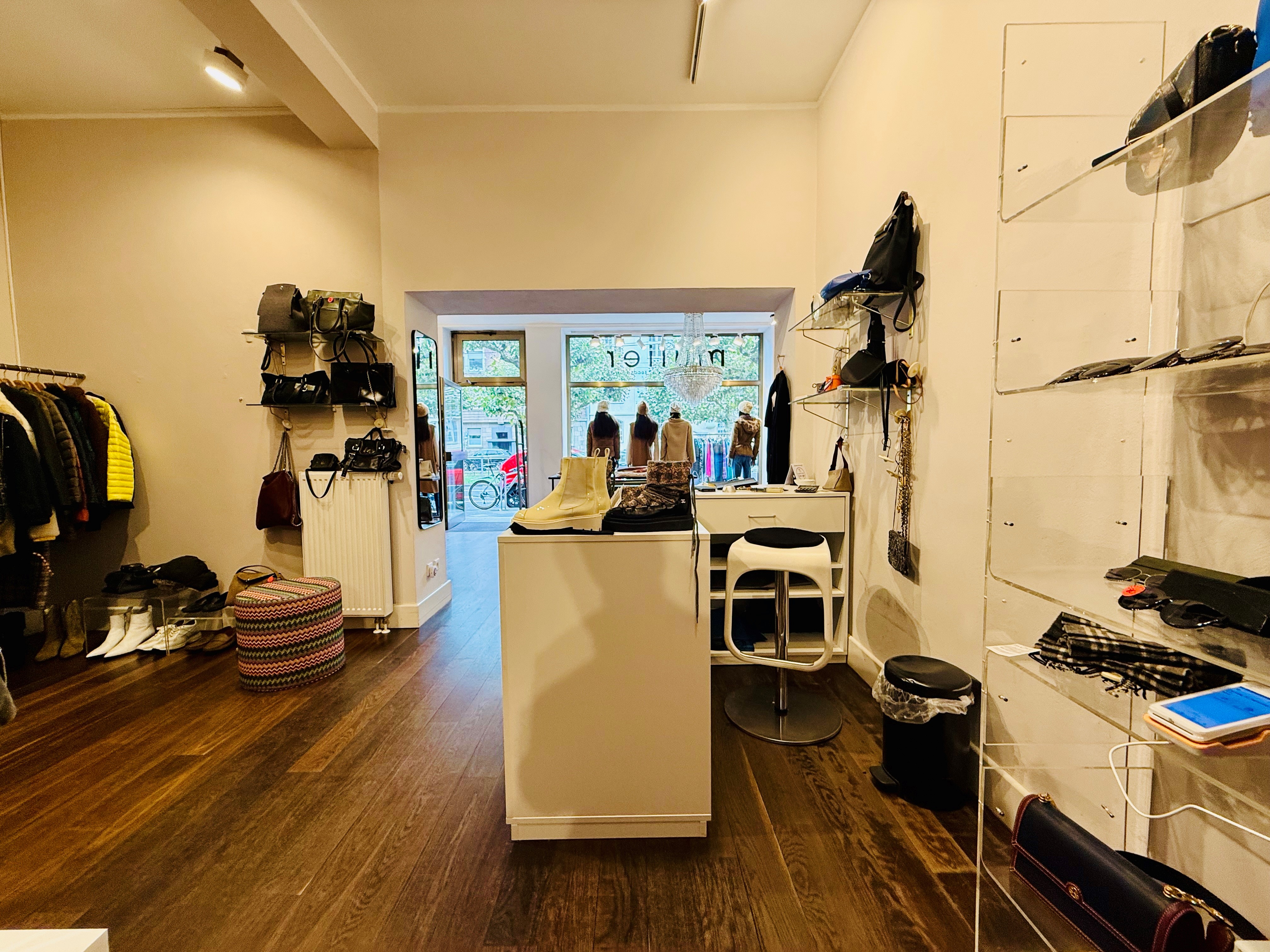 Blick aus einem Ladenlokal durch das Schaufenster nach draußen. In dem Laden hängen recht und links Textilien und wir mit einem warmen Licht beleuchtet. Der laden hat eine dunklen Holzboden.