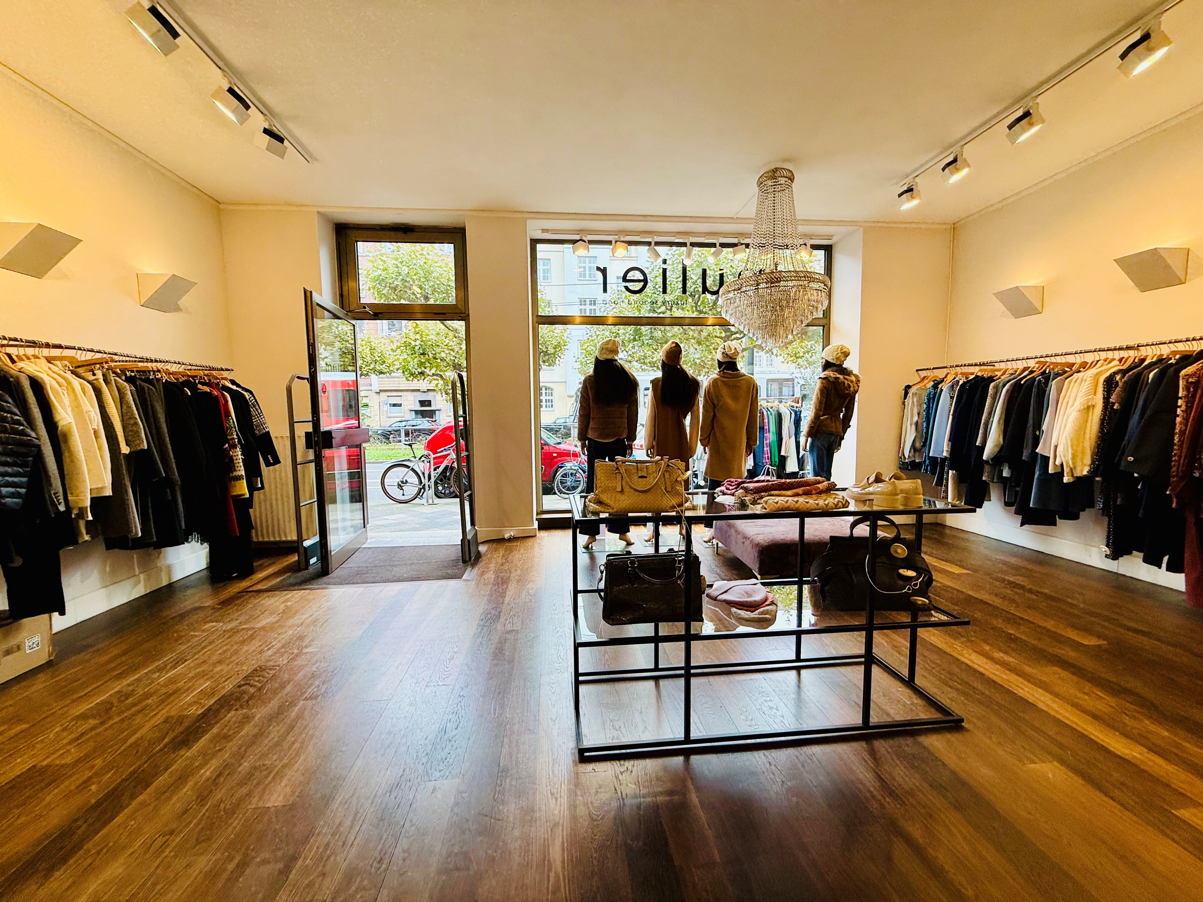 Blick aus einem Ladenlokal durch das Schaufenster nach draußen. In dem Laden hängen recht und links Textilien und wir mit einem warmen Licht beleuchtet. Der laden hat eine dunklen Holzboden.