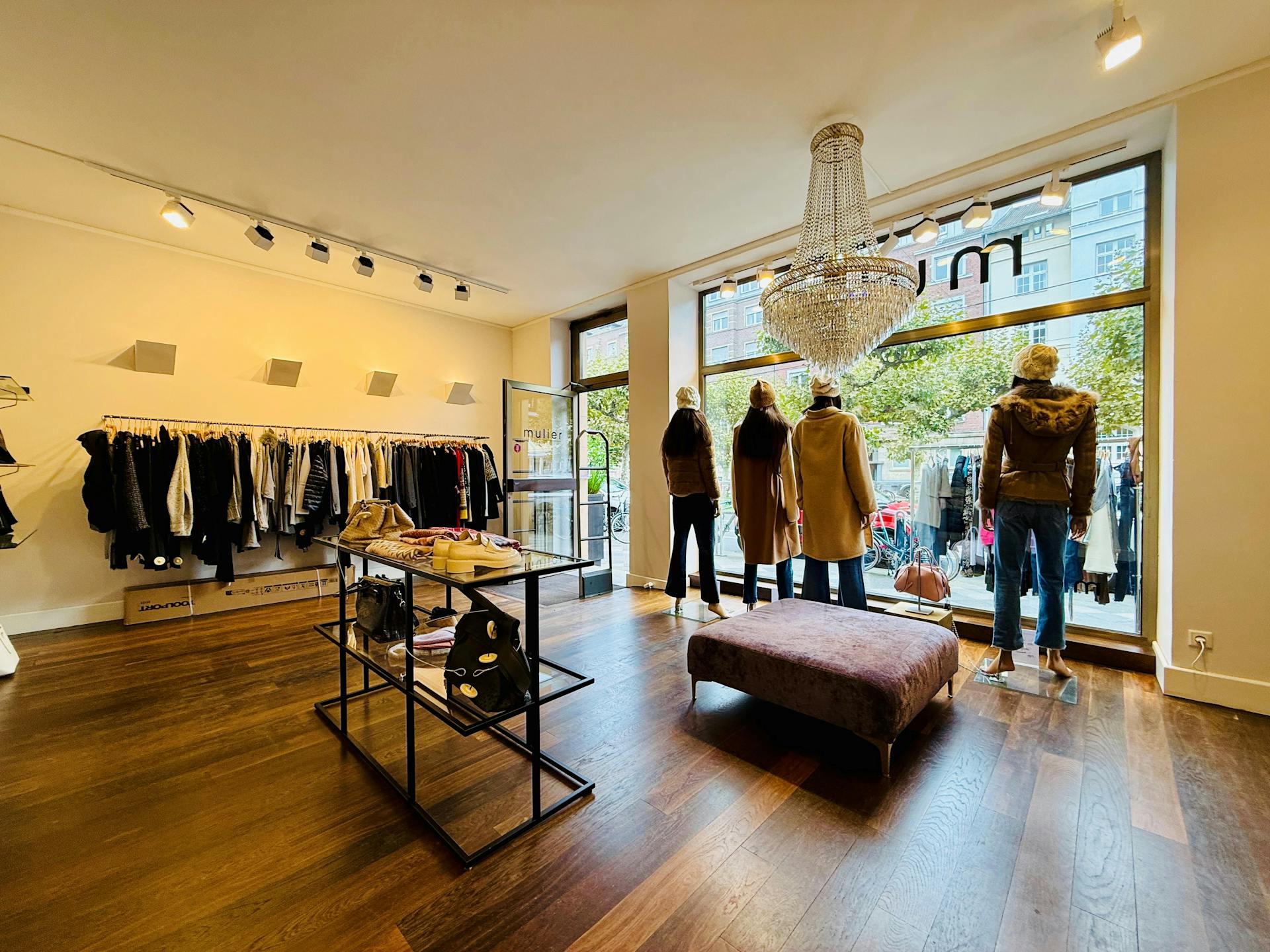 Blick aus einem Ladenlokal durch das Schaufenster nach draußen. In dem Laden hängen recht und links Textilien und wir mit einem warmen Licht beleuchtet. Der laden hat eine dunklen Holzboden.