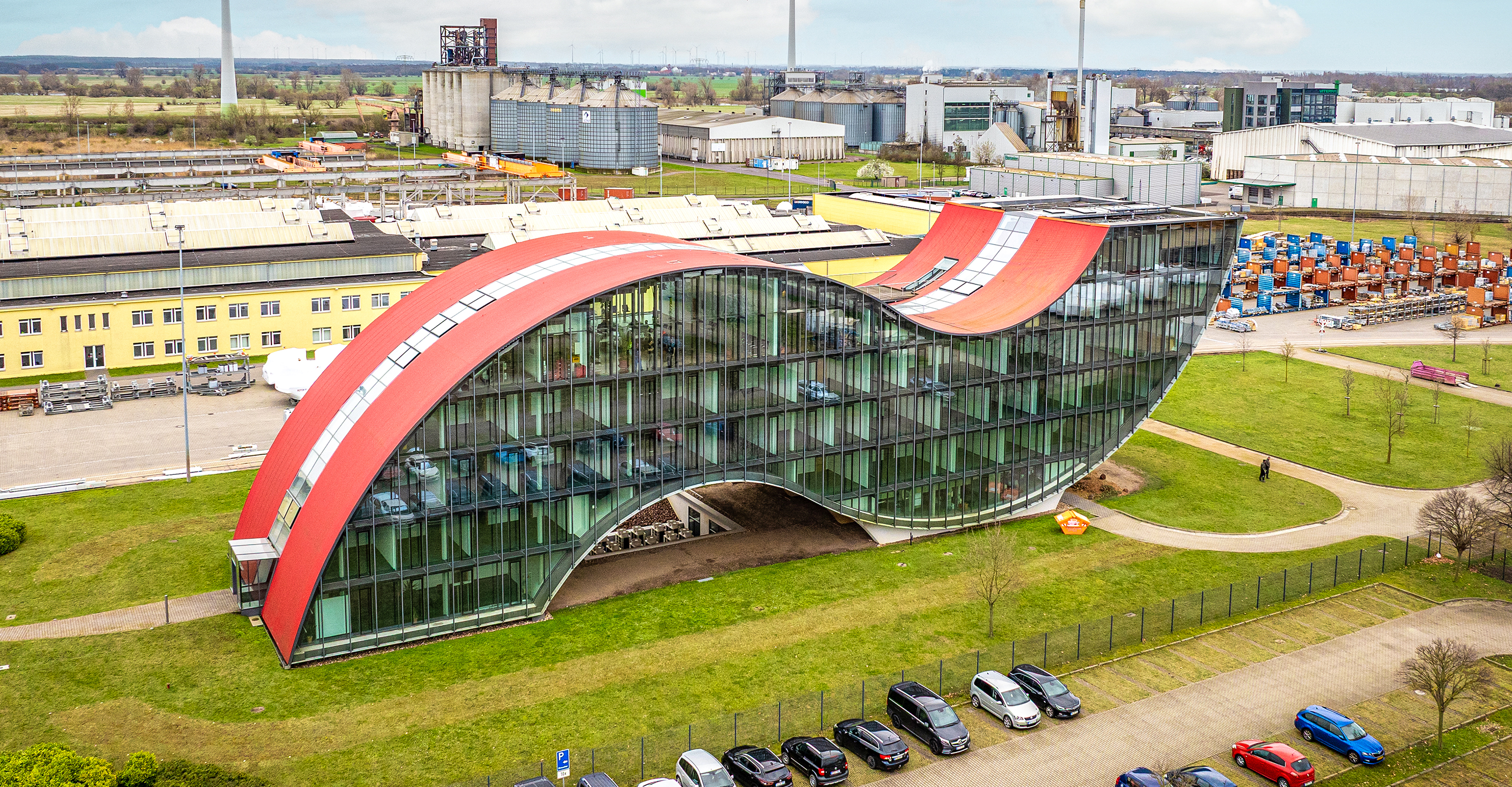 Bild eines wellenförmiges Gebäudes mit Vollverglasung und rotem Dach in Magdeburg. Im Hintergrund sind man viele Industriegebäude.