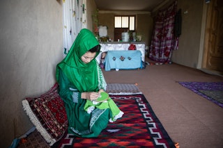 A young woman in a green hijab and dress embroiders on a carpet inside her home.