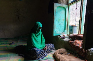 A woman in a bright green hijab looks out her window, a car battery partially obscuring her view.