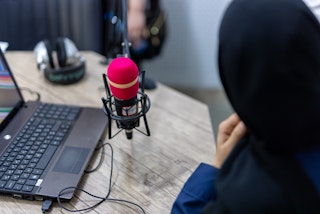 A woman radio announcer in a black hijab speaks over a red microphone