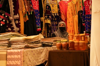 A masked woman sits in a market stall, her wares in the foreground.